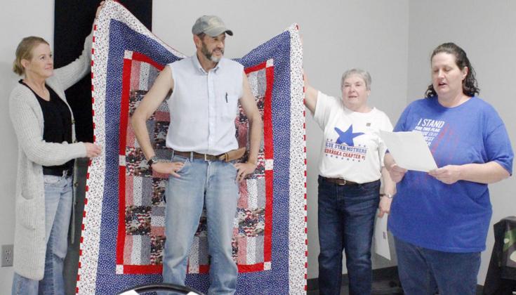 JOHN BAUM was presented with a Quilt of Valor by the Blue Star Mothers of Nebraska and the Johnson County American Legion Auxiliary. Holding the quilt up are Bobbi Albers and Mary Ann Holland, left to right, and Erica Albers reading about the meaning of each layer of the quilt.
